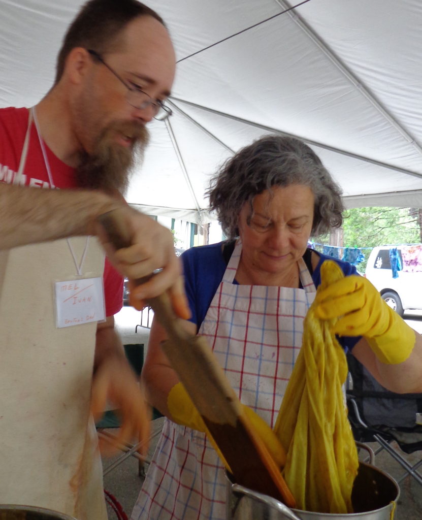 A tall white man with glasses, mustache assists a shorter woman putting yellow yarn into a dyepot. Both are wearing aprons.